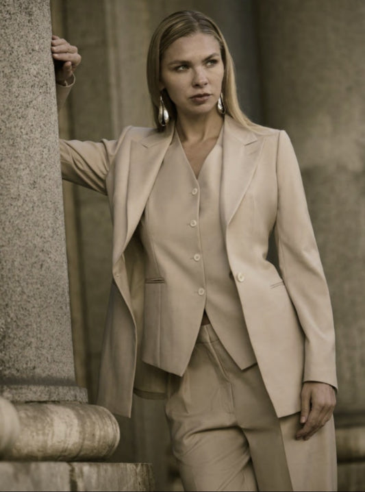 A woman wearing a beige single-breasted, three-piece trouser suit with a wide peaked lapel and straight flapless pockets.