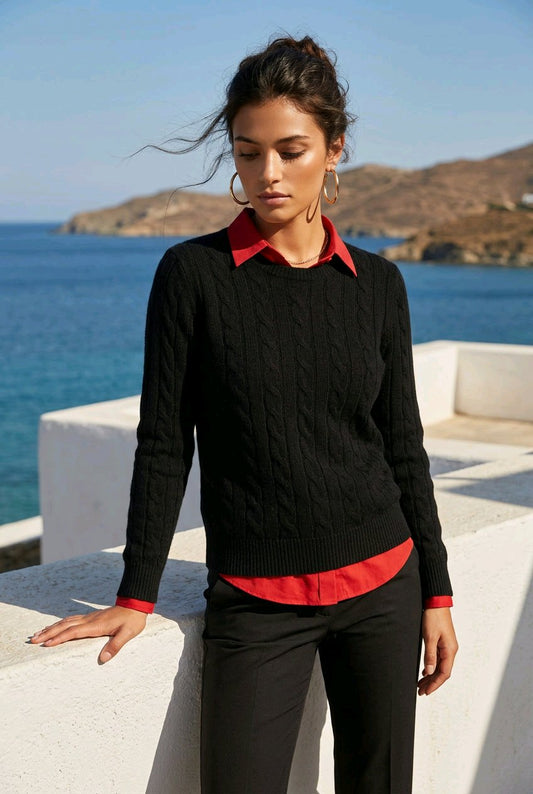 A woman wearing a black cable-knit sweater over a red collared shirt and black trousers, standing outdoors by the sea with white walls in the foreground.