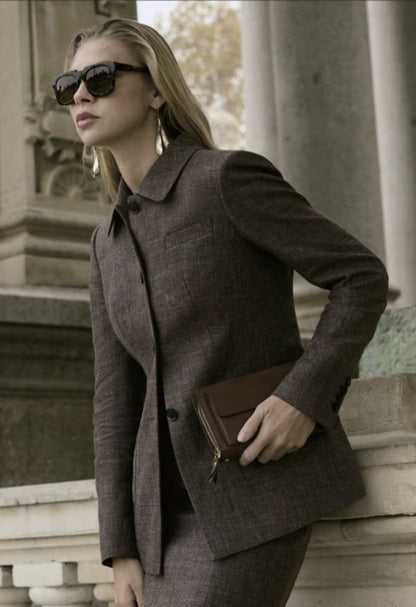 A woman wearing a brown single-breasted two-piece skirt suit with a jacket and sunglasses, standing outdoors.