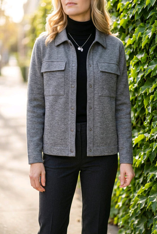 A woman wearing a gray button-front overshirt jacket over a black turtleneck and black pants, standing outdoors next to a leafy green wall.