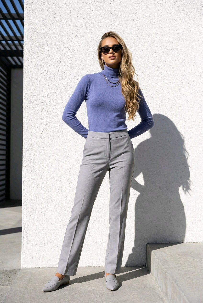 A woman wearing a lavender long-sleeve mock neck top, light gray straight-leg trousers, and gray loafers stands against a white textured wall.