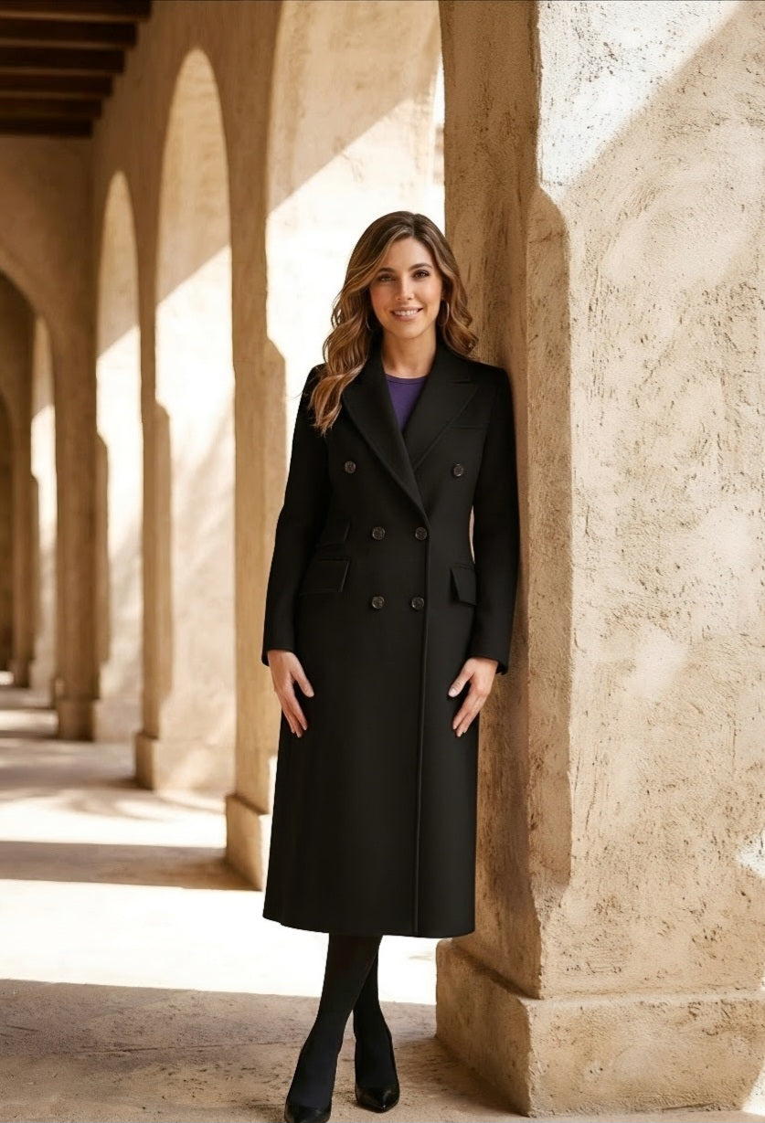 A woman wearing a long black double-breasted coat over a purple top, standing in a sunlit stone corridor with arches.