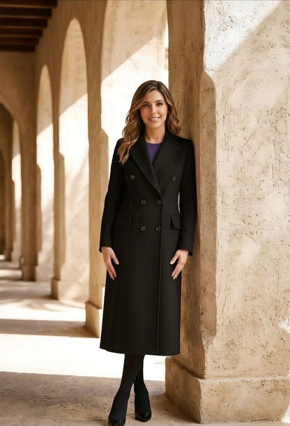 A woman wearing a long black double-breasted coat over a purple top, standing in a sunlit stone corridor with arches.
