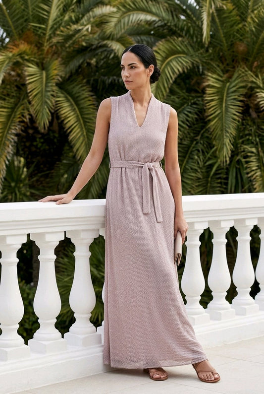 A woman wearing a sleeveless, V-neck maxi dress in a dusty rose pink color stands on a white balcony with palm trees in the background.