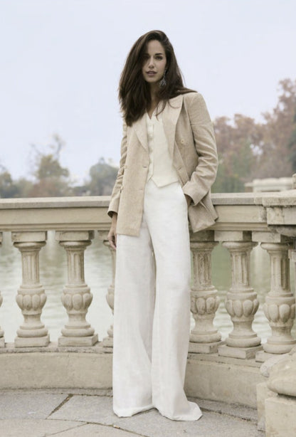 A woman wearing a tan single-breasted jacket with ivory buttons, paired with an ivory waistcoat and white trousers, standing by a balustrade.