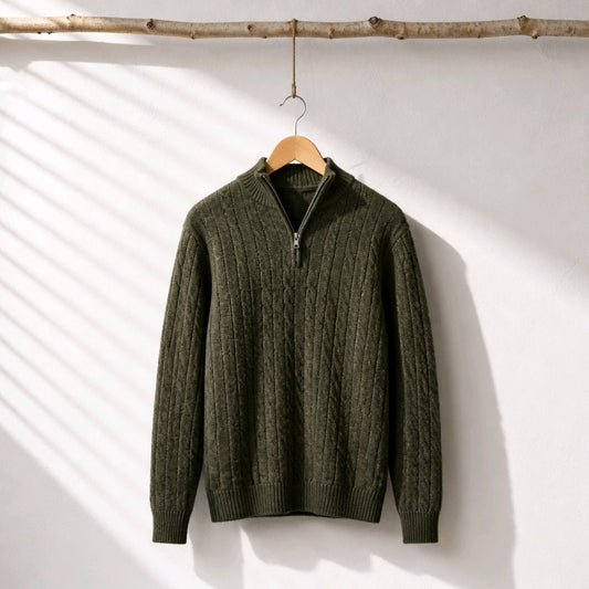 An olive green cable-knit quarter-zip sweater hanging on a wooden hanger from a rustic branch, set against a white wall with soft diagonal shadow patterns.