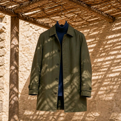 An olive green trench coat hanging on a wooden hanger against a rustic beige stucco wall under a thatched roof, with dappled sunlight and shadows across the scene.