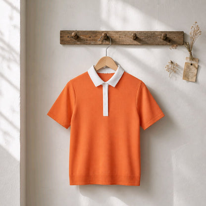 An orange short-sleeve polo shirt with a white collar and white placket, hanging on a wooden hanger from a rustic wooden peg rack against a textured off-white wall.