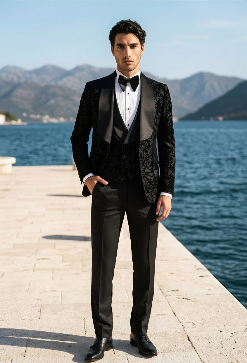 A man wearing a black tuxedo with patterned satin lapels, a white dress shirt, and a black bow tie, standing on a stone pier with a blue sea and mountains in the background.