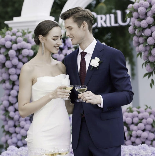 Couple in wedding attire holding glasses of champagne with a floral backdrop