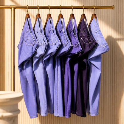 Five pairs of folded denim jeans in shades of blue and purple hang on wooden hangers from a gold clothing rack against a beige ribbed wall.