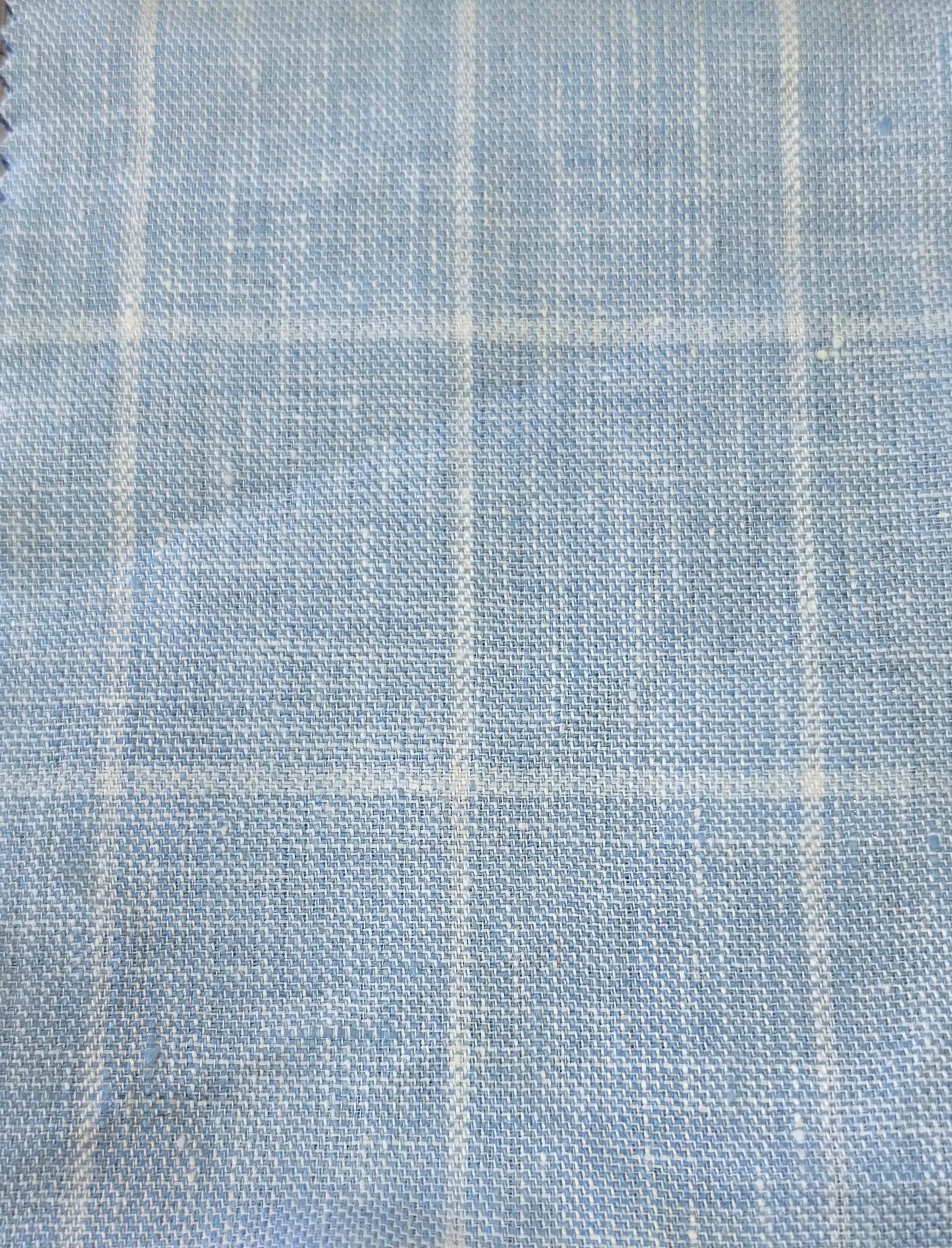 A close-up of light blue woven fabric with a subtle white windowpane plaid pattern.