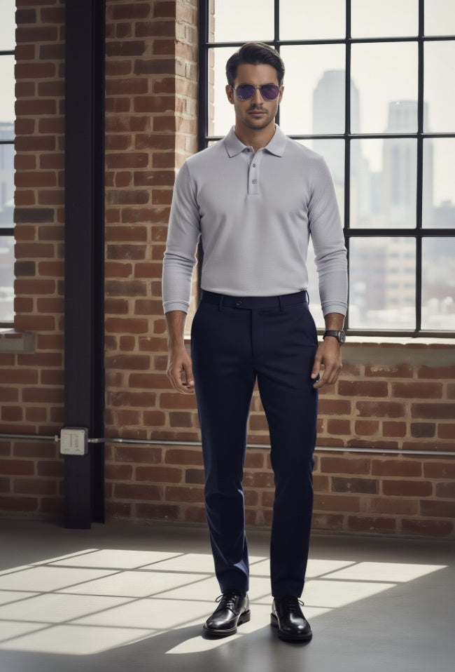 Man wearing a light gray long-sleeve polo shirt and dark blue pants standing in front of a brick wall with large windows.