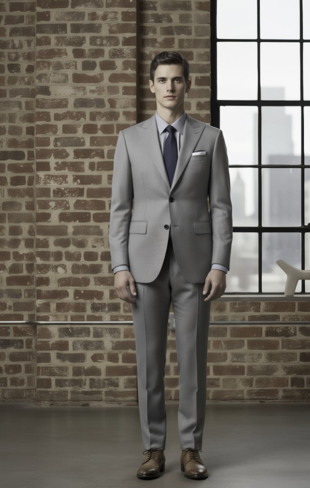 Man wearing a light grey suit standing against a brick wall with a window in the background