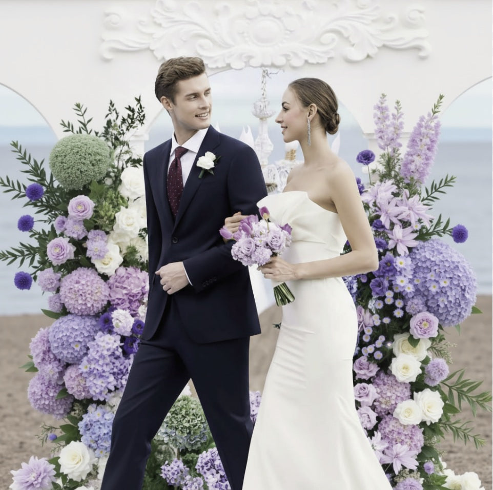 Man and woman in formal attire standing in front of a floral arch on a beach.