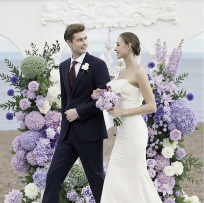 Man and woman in formal attire standing in front of a floral arch on a beach.