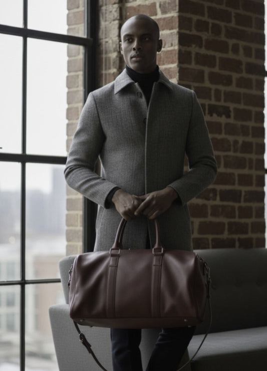Man holding a brown leather bag against a brick wall with large windows.