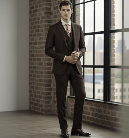 Man in a 3pc brown suit standing in a room with brick walls and large windows.