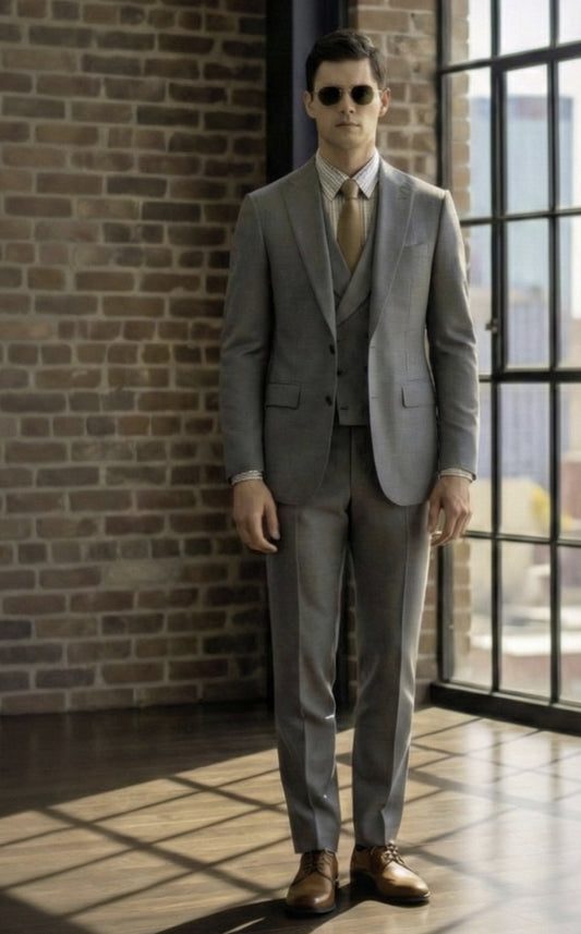 Man in a 3pc grey suit standing in a room with brick walls and large windows.