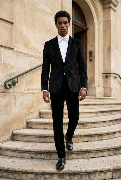 A man wearing a black textured tuxedo jacket with black trousers, a white dress shirt, and a white bow tie walks down stone steps in front of a beige stone building.
