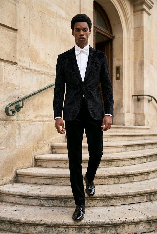 A man wearing a black textured tuxedo jacket with black trousers, a white dress shirt, and a white bow tie walks down stone steps in front of a beige stone building.