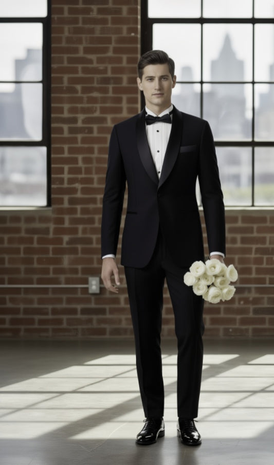Man in a black tuxedo holding a bouquet of flowers in a room with large windows and brick walls.