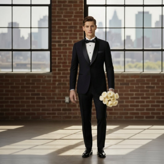 Man in a black tuxedo holding a bouquet of flowers in a room with large windows and brick walls.