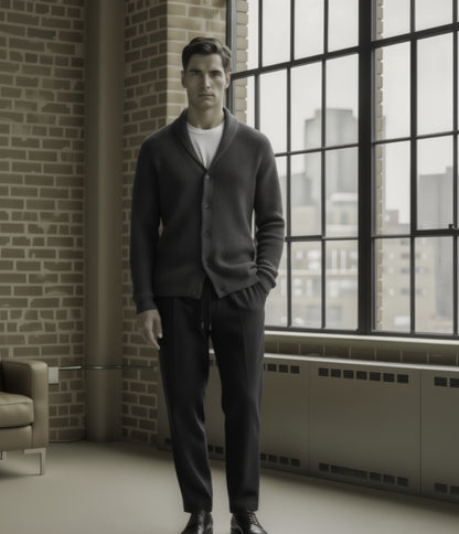 Man in a grey cardigan standing in a room with large windows and brick wall.