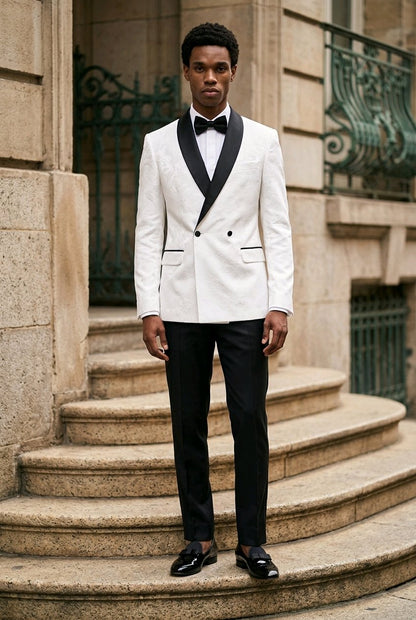 A man wearing a white double-breasted tuxedo jacket with black satin lapels, black trousers, a black bow tie, and black dress shoes, standing on stone steps in front of a building.