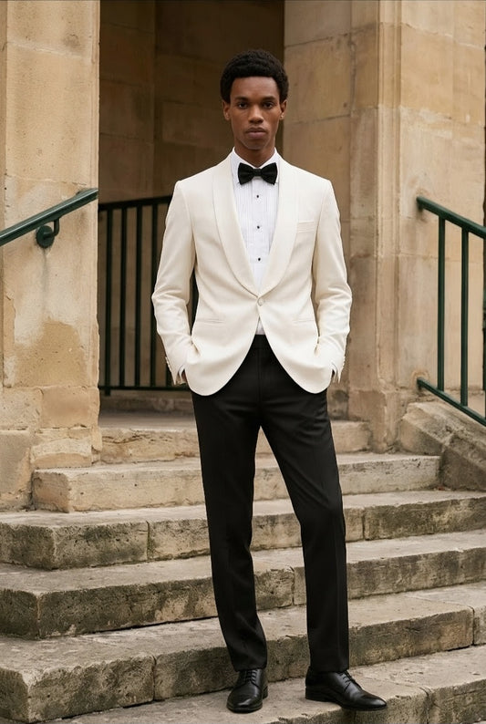 A man wearing a white single-breasted tuxedo jacket with black trousers, a white shirt, and a black bow tie, standing on stone steps in front of a stone building.
