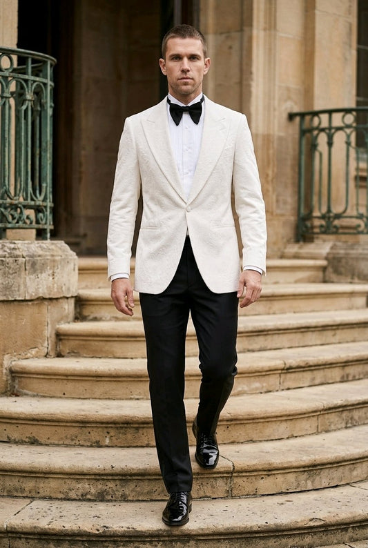 A man wearing a white textured tuxedo jacket with black trousers, a white shirt, and a black bow tie walks down stone steps in front of a classical building.