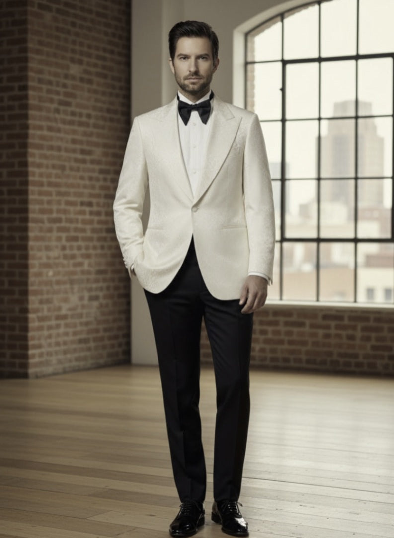 Man in a white tuxedo with black bow tie standing in a room with brick walls and large windows.