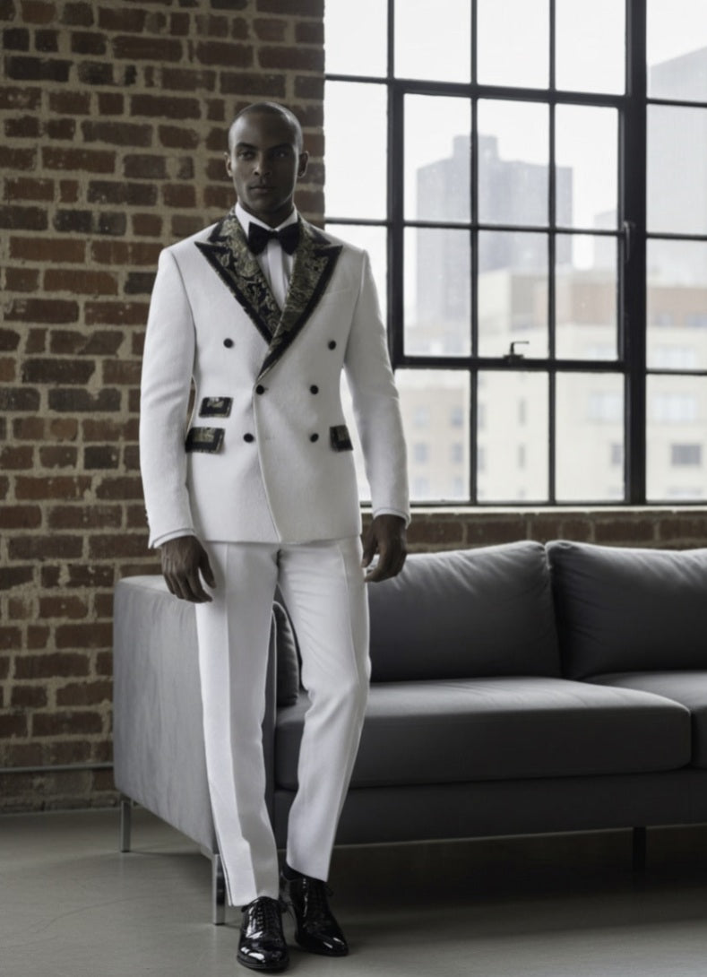 Man in a white tuxedo with black lapels standing in a room with a brick wall and large window.