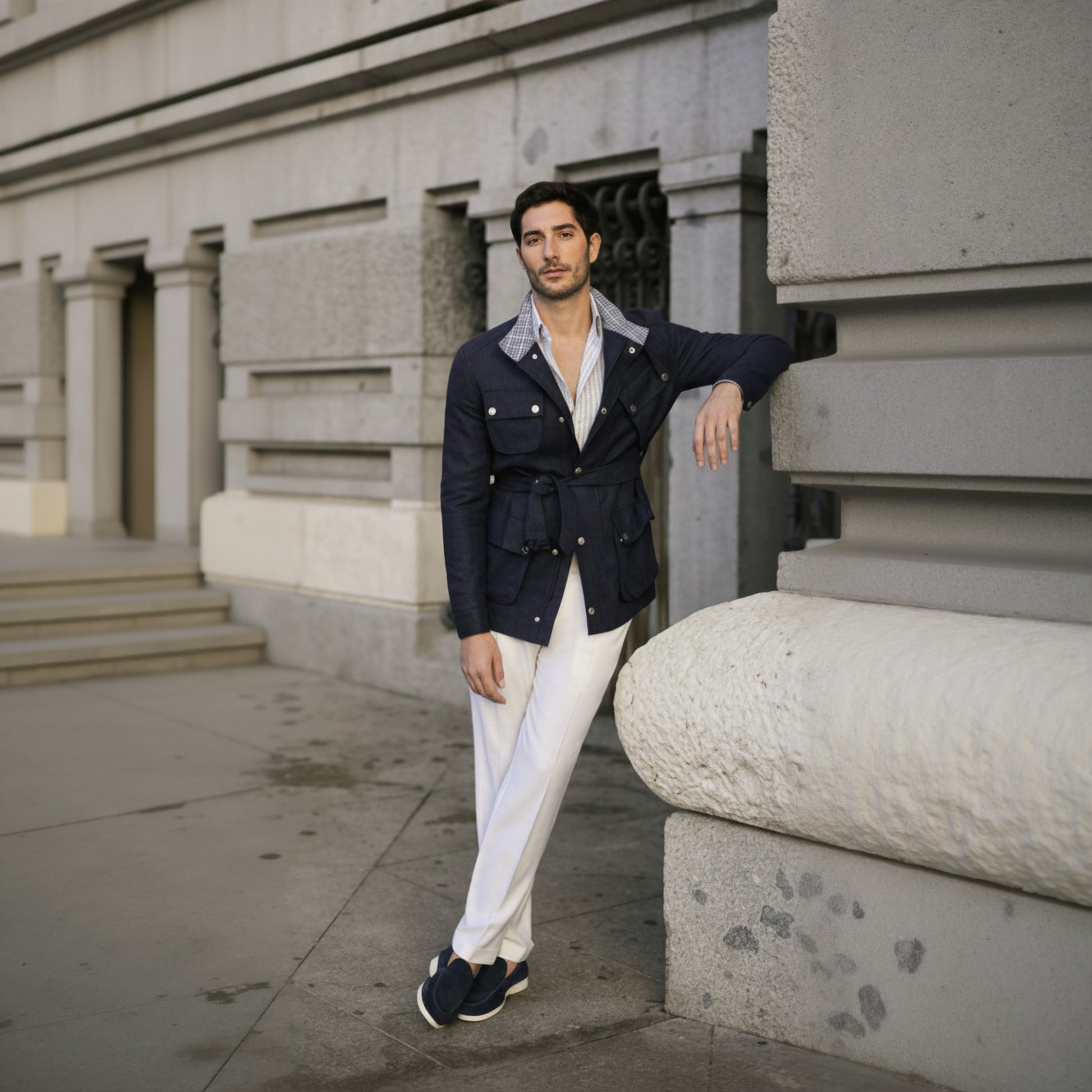 Man in navy jacket and white pants leaning against a stone building.