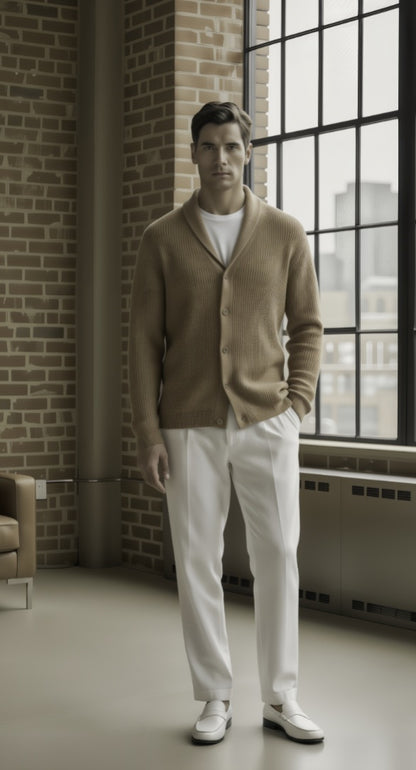 Man wearing a brown cardigan over a white shirt and pants, standing in a room with brick walls and large windows.