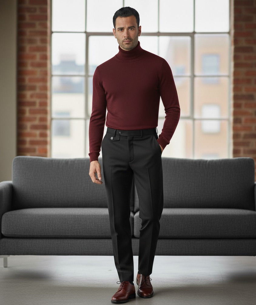 Man wearing a burgundy turtleneck sweater and dark pants standing in a room with a gray couch and large windows.