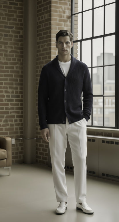 Man wearing a dark cardigan, white shirt, and white pants standing in a room with brick walls and large windows.