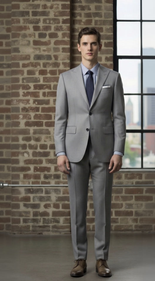 Man wearing a light grey suit standing against a brick wall with a window in the background