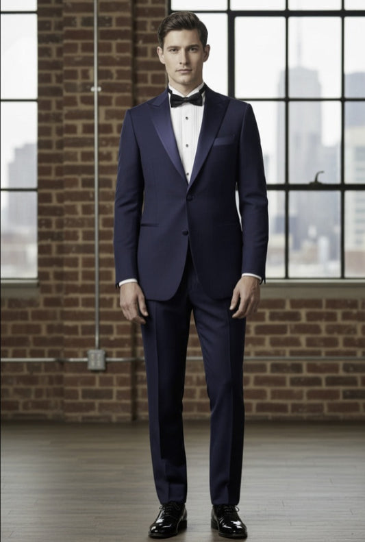 Man wearing a navy blue tuxedo with a white shirt and black bow tie, standing in front of a brick wall.