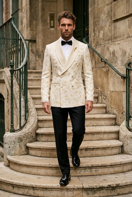A man wearing a cream-colored double-breasted tuxedo jacket with gold damask embroidery, black trousers, and a black bow tie walks down stone stairs in an outdoor setting.