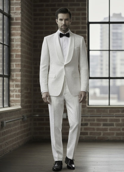 Man wearing a white tuxedo with a bow tie in an urban setting with brick walls and large windows.