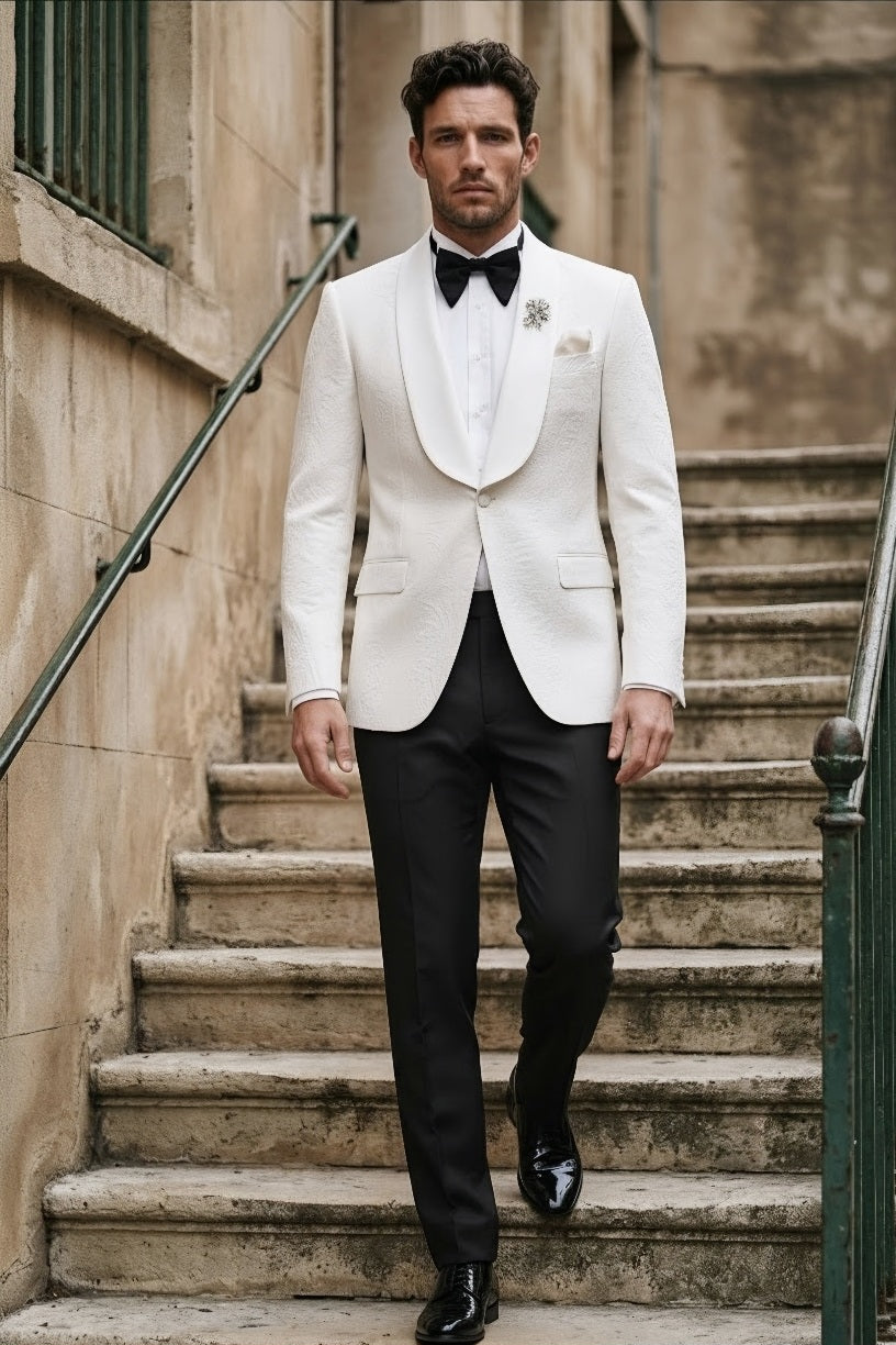 A man walks down outdoor stone stairs wearing a white tuxedo jacket with black trousers, a white shirt, a black bow tie, and a black dress shoe.