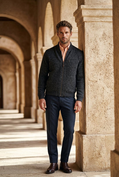 A man stands in a stone colonnade wearing a dark charcoal zip-front knit jacket over a peach shirt, dark navy trousers, and brown leather shoes.
