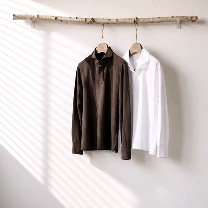 Two long-sleeve collared shirts hanging on a natural wood branch rack against a white wall with soft diagonal shadow stripes. The left shirt is dark brown and the right shirt is white.