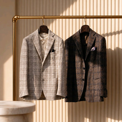 Two tailored blazers hanging on a brass clothing rack against a beige ribbed wall. The left blazer is light beige with a subtle plaid pattern, and the right blazer is dark brown with a darker plaid pattern.