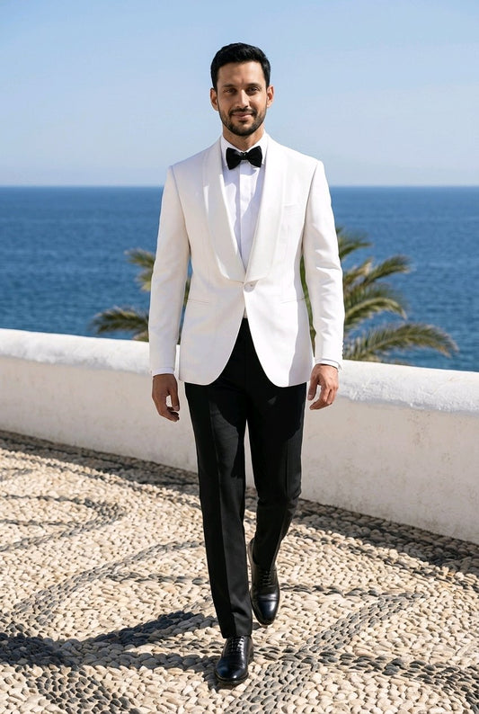 A man wearing a white tuxedo jacket with black peak lapels, a white dress shirt, a black bow tie, and black trousers, walking outdoors with a sea and sky background.