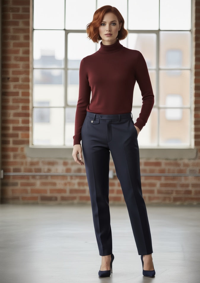 Woman wearing a burgundy turtleneck and dark pants standing in front of a brick wall with large windows.