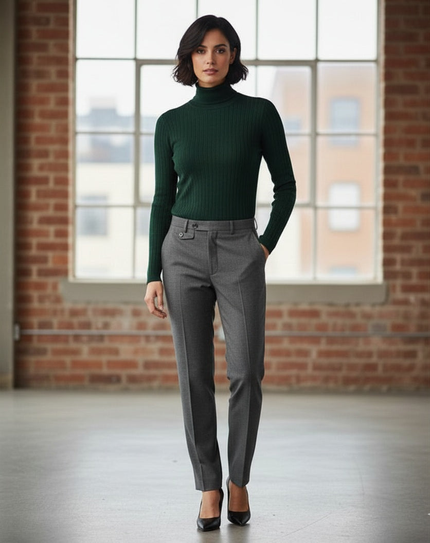 Woman wearing a green turtleneck and gray pants standing in front of a brick wall with large windows.
