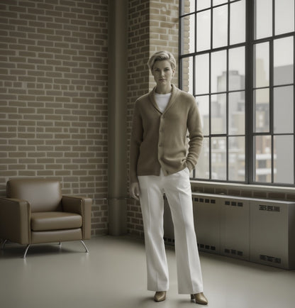 Women standing in a room with a brick wall and large window, wearing a beige cardigan and white pants.