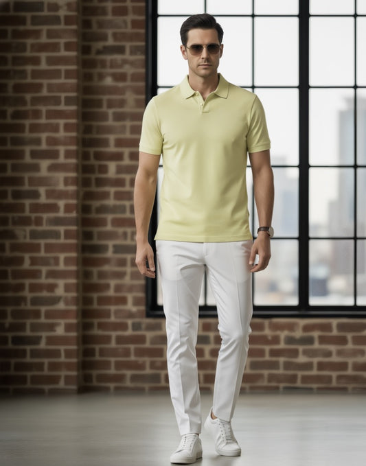 Man wearing a light yellow polo shirt and white pants standing against a brick wall.